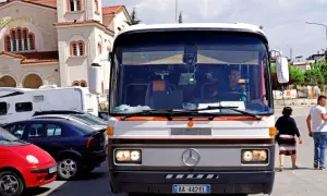 Autobus w Albanii, z kierowcą za kierownicą, na tle kościoła. W tle widać też inne pojazdy.