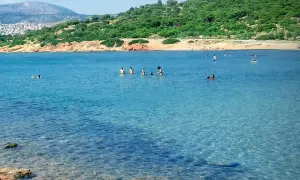 Słoneczna **Ateny plaża** z turkusową wodą, gdzie ludzie kąpią się i relaksują. W tle zielone wzgórza i zabudowania.