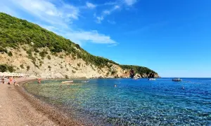 Kamienista plaża z turkusową wodą, zielonymi wzgórzami i kilkoma łodziami na morzu.