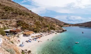 Słoneczna plaża w Porto Palermo z turkusową wodą, leżakami i łódkami.