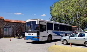 Biały macedoński autobus z niebieskimi pasami stoi na parkingu obok kamiennego muru i drzew.