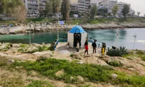 Mała kapliczka nad wodą, z grecką flagą, na tle budynków. Widok na pireus plaża.