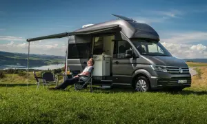 Mężczyzna odpoczywa na krześle obok szarego VW Transporter kampera, rozkoszując się widokiem jeziora i wzgórz.