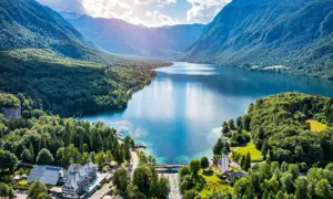 Malownicze jezioro Bohinj otoczone zielonymi wzgórzami i górami. Widać most, kościół i hotel.