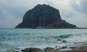 Monemvasia, Grecja: skalista góra z ruinami zamku na szczycie, jacht na turkusowym morzu i kamienista plaża.