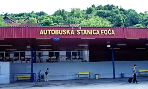 Autobuska stanica Foča, zalesione wzgórza w tle. Mężczyzna z walizką na kółkach, dzieci idące obok. Marka konwertybilna.
