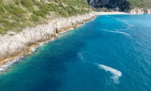 Malownicze wybrzeże z zielonymi wzgórzami i skalistymi klifami schodzącymi do turkusowego morza. W oddali widać piaszczystą plażę Gjipe.