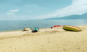 Spokojny brzeg Jeziora Ochrydzkiego w Albanii. Kilka łódek, w tym kolorowa łódka i czerwona rowerowa, odpoczywa na piaszczystej plaży.