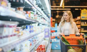 Kobieta z wózkiem pełnym warzyw i owoców w supermarkecie. Sprawdza rumunia ceny w sklepach, porównując produkty na półkach.