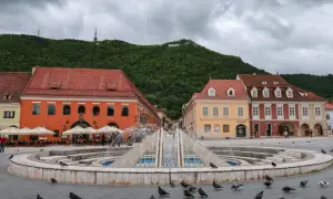 Fontanna na placu w Braszowie, z kolorowymi kamienicami i napisem "BRASOV" na wzgórzu. Wiele gołębi spaceruje wokół.