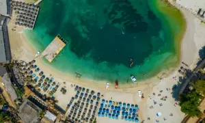 Widok z lotu ptaka na turkusowe morze i piaszczystą plażę z rzędami leżaków i parasoli. Woda jest krystalicznie czysta, widać ludzi pływających i łodzie. To idealne miejsce na relaks na **tirana plaże**.