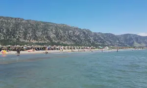 Słoneczny dzień na plaży Velipoja. Ludzie odpoczywają pod parasolami, kąpią się w morzu, a w tle wznoszą się górzyste tereny.