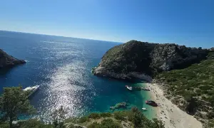 Słoneczna zatoka grama z turkusową wodą, kamienistą plażą pełną ludzi i łodziami przycumowanymi do brzegu.