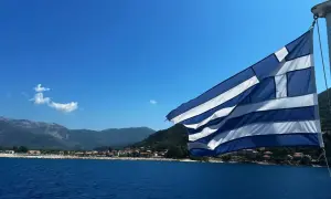 Flaga Grecji powiewa nad błękitnym morzem, w tle widać górzyste wybrzeże z zabudowaniami. Idealny dzień na plażowanie w Grecji.