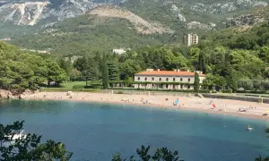Malownicza zatoka z plażą w Czarnogórze. Woda jest turkusowa, a plaża piaszczysta, z kilkoma osobami odpoczywającymi pod parasolami. W tle widać zielone wzgórza i elegancki budynek.