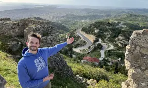 Mężczyzna na tle malowniczego krajobrazu z krętą drogą i miastem w oddali. To piękna Albania zimą, choć zieleń sugeruje wczesną wiosnę.
