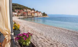 Kamienista plaża z widokiem na kolorowe domki na wzgórzu. To właśnie takie widoki sprawiają, że Istria ma najpiękniejsze plaże.
