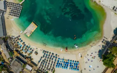 Widok z lotu ptaka na turkusowe morze i piaszczystą plażę z rzędami leżaków i parasoli. Woda jest krystalicznie czysta, widać ludzi pływających i łodzie. To idealne miejsce na relaks na **tirana plaże**.