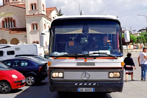 Autobus w Albanii, z kierowcą za kierownicą, na tle kościoła. W tle widać też inne pojazdy.