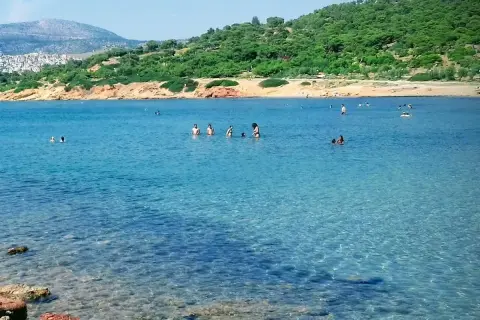 Słoneczna **Ateny plaża** z turkusową wodą, gdzie ludzie kąpią się i relaksują. W tle zielone wzgórza i zabudowania.