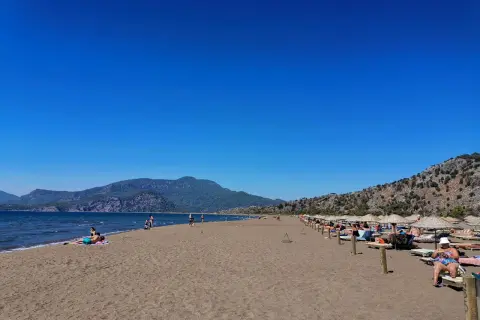 Plaża żółwi w Turcji, z turkusowym morzem i górami w tle. Ludzie odpoczywają na leżakach pod parasolami.