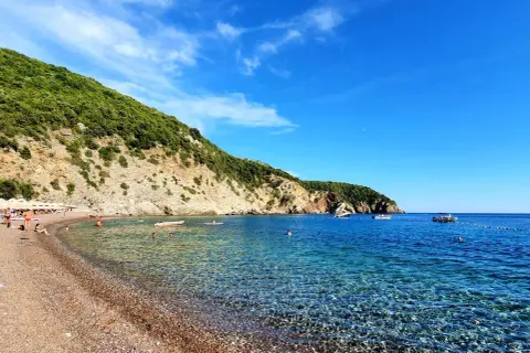 Kamienista plaża z turkusową wodą, zielonymi wzgórzami i kilkoma łodziami na morzu.