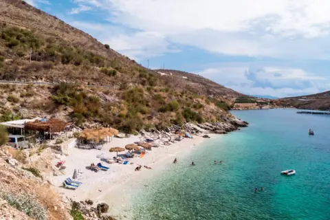 Słoneczna plaża w Porto Palermo z turkusową wodą, leżakami i łódkami.