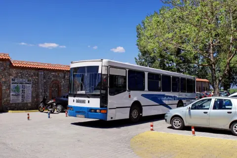 Biały macedoński autobus z niebieskimi pasami stoi na parkingu obok kamiennego muru i drzew.