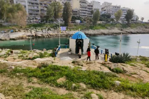 Mała kapliczka nad wodą, z grecką flagą, na tle budynków. Widok na pireus plaża.