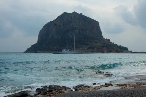 Monemvasia, Grecja: skalista góra z ruinami zamku na szczycie, jacht na turkusowym morzu i kamienista plaża.