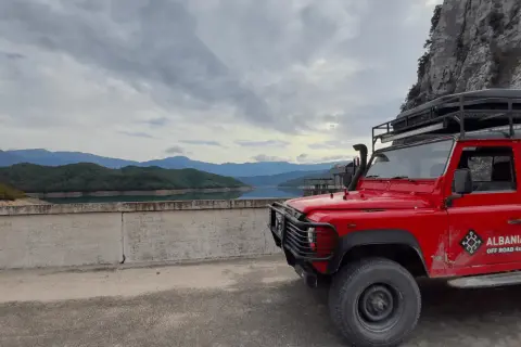 Czerwony Land Rover na tle gór i jeziora. Chłodna, pochmurna pogoda, typowa dla Albanii w styczniu.