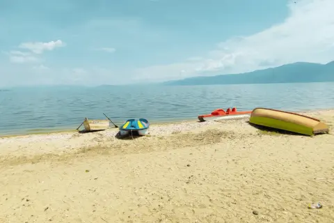 Spokojny brzeg Jeziora Ochrydzkiego w Albanii. Kilka łódek, w tym kolorowa łódka i czerwona rowerowa, odpoczywa na piaszczystej plaży.