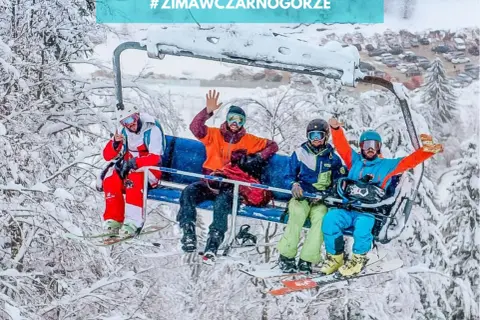 Czterech narciarzy na wyciągu krzesełkowym cieszy się zimą w Czarnogórze. W tle zaśnieżone drzewa i parking.