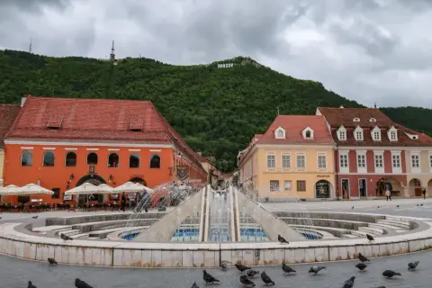 Fontanna na placu w Braszowie, z kolorowymi kamienicami i napisem "BRASOV" na wzgórzu. Wiele gołębi spaceruje wokół.