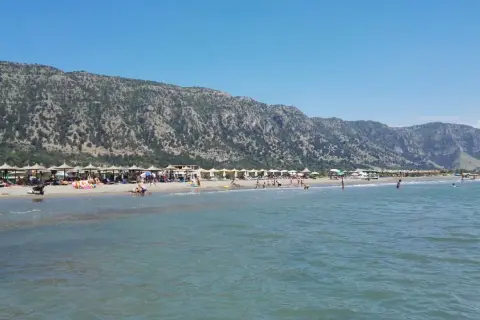 Słoneczny dzień na plaży Velipoja. Ludzie odpoczywają pod parasolami, kąpią się w morzu, a w tle wznoszą się górzyste tereny.