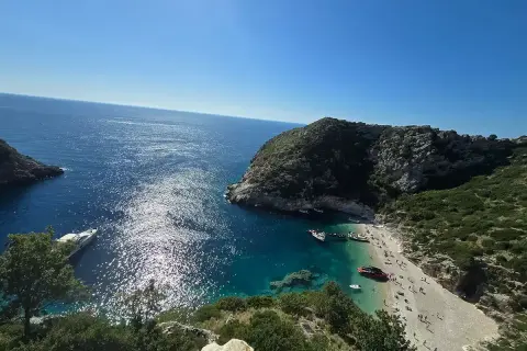 Słoneczna zatoka grama z turkusową wodą, kamienistą plażą pełną ludzi i łodziami przycumowanymi do brzegu.