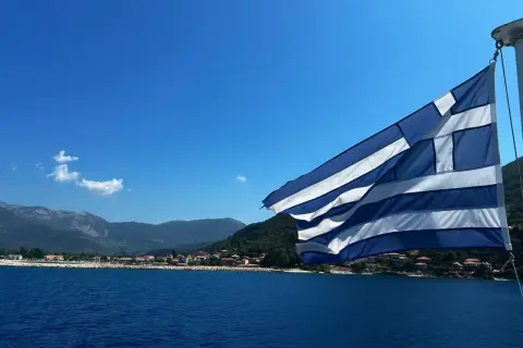 Flaga Grecji powiewa nad błękitnym morzem, w tle widać górzyste wybrzeże z zabudowaniami. Idealny dzień na plażowanie w Grecji.