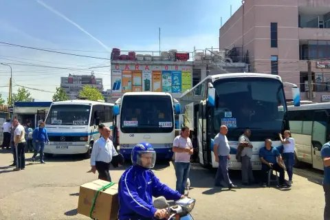Autobusy na dworcu, jeden z nich jedzie do Vlory, inny do Tirany. Ludzie czekają na podróż.