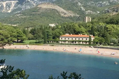 Malownicza zatoka z plażą w Czarnogórze. Woda jest turkusowa, a plaża piaszczysta, z kilkoma osobami odpoczywającymi pod parasolami. W tle widać zielone wzgórza i elegancki budynek.