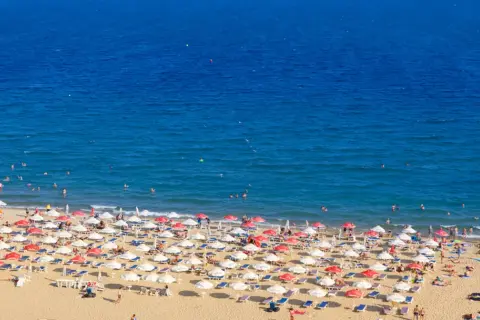 Słoneczny brzeg tętni życiem. Tłumy plażowiczów odpoczywają pod parasolami, kąpią się w morzu i opalają na piasku.