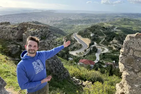 Mężczyzna na tle malowniczego krajobrazu z krętą drogą i miastem w oddali. To piękna Albania zimą, choć zieleń sugeruje wczesną wiosnę.