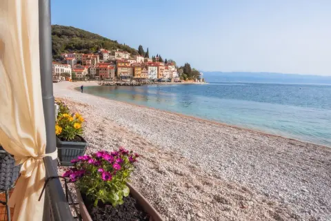 Kamienista plaża z widokiem na kolorowe domki na wzgórzu. To właśnie takie widoki sprawiają, że Istria ma najpiękniejsze plaże.