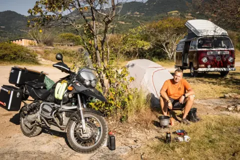 Motocykl, namiot i kamper to idealny zestaw, by wiedzieć, co zabrać na camping. Mężczyzna gotuje na kuchence turystycznej.