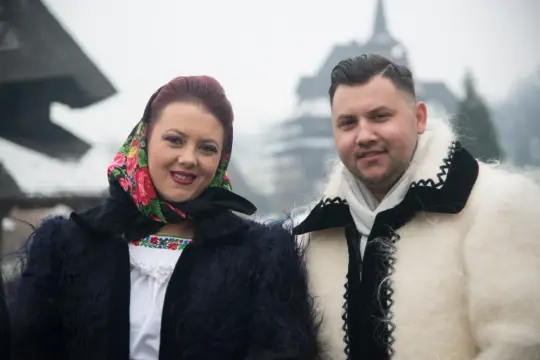 Uśmiechnięci rumuńscy ludzie w tradycyjnych strojach, kobieta w kwiecistej chuście, mężczyzna w białym futrze.
