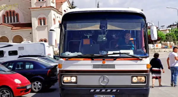 Autobus w Albanii, z kierowcą za kierownicą, na tle kościoła. W tle widać też inne pojazdy.