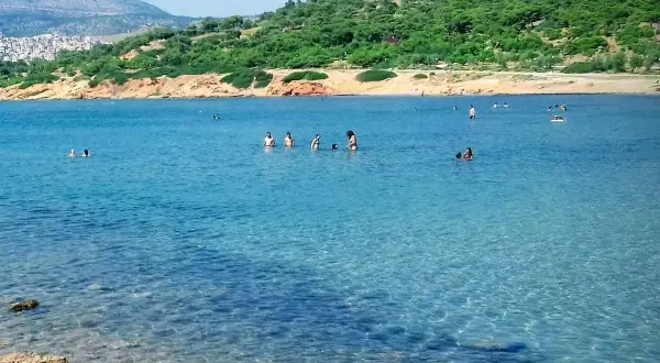 Słoneczna **Ateny plaża** z turkusową wodą, gdzie ludzie kąpią się i relaksują. W tle zielone wzgórza i zabudowania.