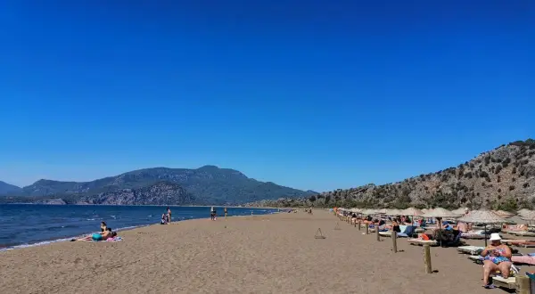 Plaża żółwi w Turcji, z turkusowym morzem i górami w tle. Ludzie odpoczywają na leżakach pod parasolami.