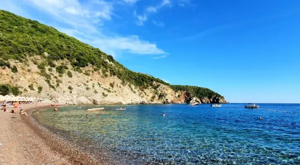 Kamienista plaża z turkusową wodą, zielonymi wzgórzami i kilkoma łodziami na morzu.