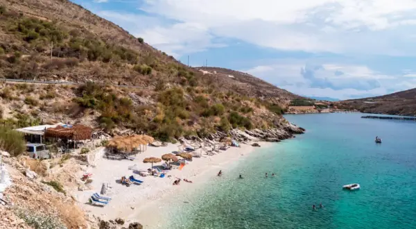 Słoneczna plaża w Porto Palermo z turkusową wodą, leżakami i łódkami.