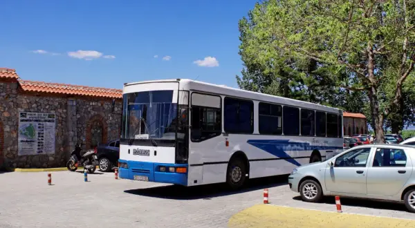 Biały macedoński autobus z niebieskimi pasami stoi na parkingu obok kamiennego muru i drzew.